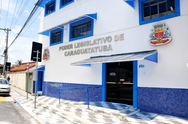 Foto mostra a fachada de um prédio institucional com a inscrição “Poder Legislativo de Caraguatatuba”, pintado em branco com detalhes em azul, com entrada principal em vidro e acessos com corrimão; o local fica à beira de uma rua, com calçada e postes ao redor, caracterizando um ambiente urbano e administrativo.
