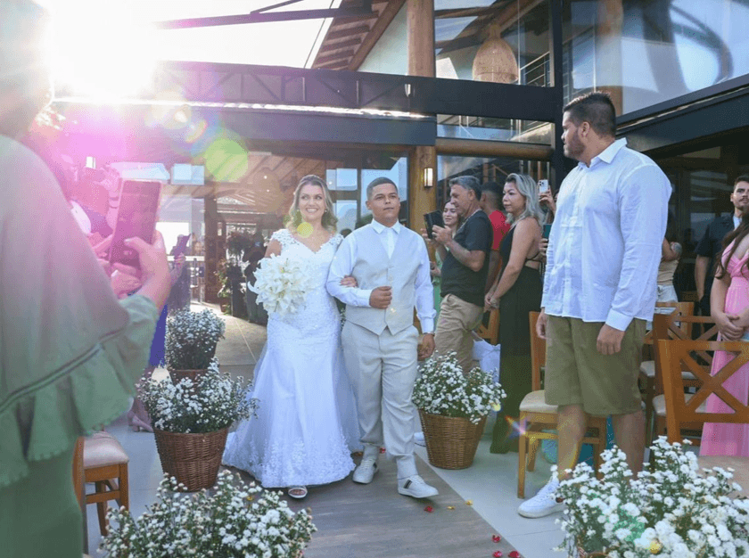 Registro da entrada de uma noiva, vestida de branco e segurando um buquê de flores, acompanhada por um jovem enquanto convidados acompanham a cerimônia.