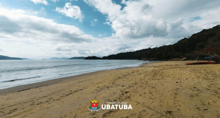 Vista ampla de uma praia tranquila em Ubatuba, com faixa de areia extensa e quase vazia, mar calmo e céu parcialmente encoberto por nuvens. Ao fundo, a vegetação da Mata Atlântica cobre as encostas próximas ao litoral, criando uma paisagem natural e serena.