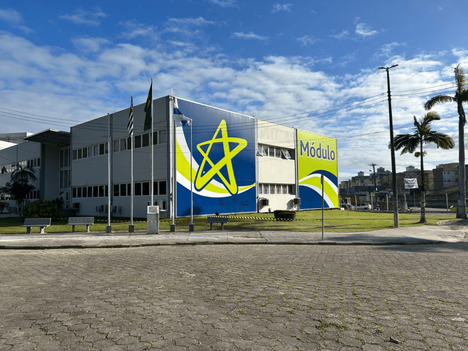 Fachada de um edifício moderno identificado como “Módulo”, com pintura nas cores azul, verde e branco e um grande símbolo em formato de estrela na lateral. Em frente ao prédio, há mastros com bandeiras, bancos, gramado e algumas palmeiras, sob um céu azul com nuvens espalhadas.