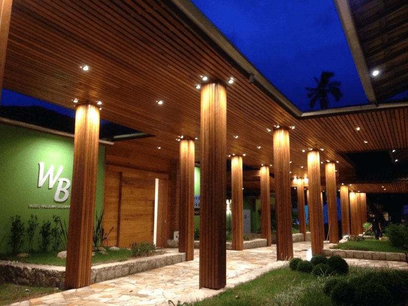 Entrada de um edifício com arquitetura moderna, destacada por colunas revestidas de madeira, teto com ripas e iluminação embutida. Um caminho de pedras atravessa jardins bem cuidados até a porta principal, enquanto o céu ao entardecer cria um contraste com a iluminação quente do local.