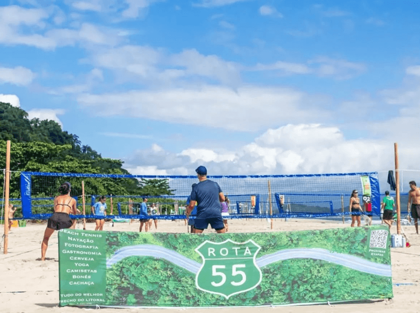 Pessoas praticam vôlei de praia em quadras de areia à beira-mar, sob um céu azul com nuvens. Em primeiro plano há uma placa com a marca "Rota 55", enquanto ao fundo aparecem outras quadras, atletas em atividade e a vegetação da encosta litorânea.