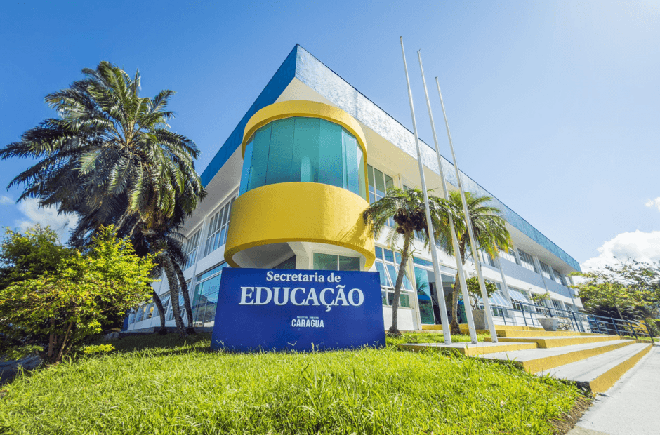 Fachada da Secretaria de Educação de Caraguatatuba, um prédio moderno de dois andares com detalhes nas cores azul, branco e amarelo. Em frente ao edifício há um gramado, palmeiras, mastros para bandeiras e uma placa azul com o nome da secretaria, sob um céu claro e ensolarado.