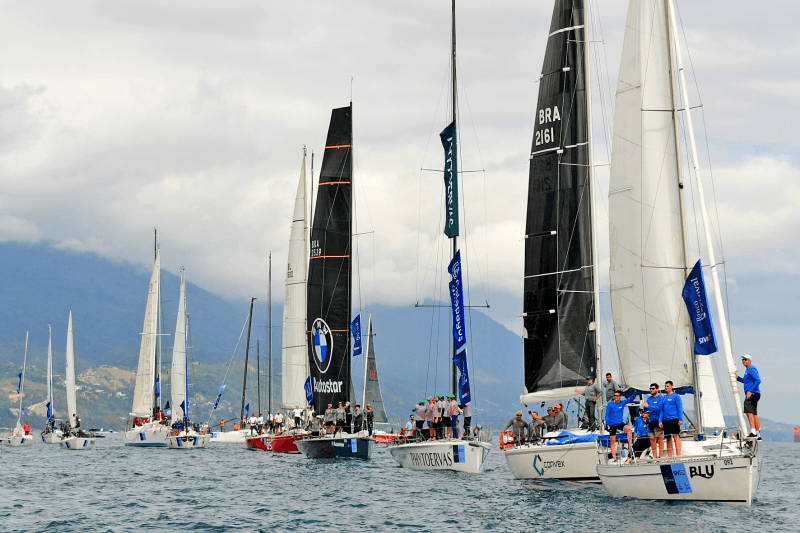 Regata de veleiros em mar aberto, com diversas embarcações alinhadas sobre a água. As velas brancas e pretas se destacam diante do céu nublado e das montanhas ao fundo, enquanto equipes de tripulantes permanecem a bordo, compondo uma cena que celebra o esporte náutico e a paisagem litorânea.
