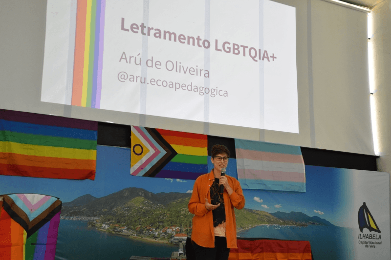 Em um auditório, uma pessoa segura um microfone e faz uma apresentação em frente a um telão com o título “Letramento LGBTQIA+”. Ao fundo, bandeiras com as cores do arco-íris e da comunidade trans decoram o espaço, junto a um painel com imagem de paisagem litorânea e a identificação de Ilhabela.