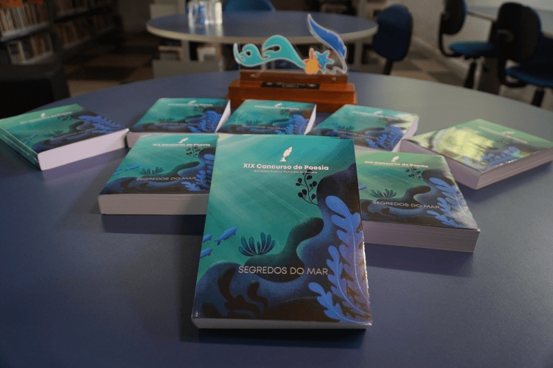 Em uma mesa de biblioteca, vários exemplares do livro Segredos do Mar, do XIX Concurso de Poesia, estão dispostos ao redor de um troféu. As capas em tons de azul e verde trazem ilustrações inspiradas no fundo do mar, enquanto o ambiente ao fundo apresenta mesas, cadeiras e estantes de livros desfocadas.